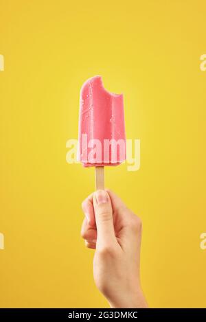 Woman holds bitten red ice cream on yellow background. Color frozen fruit popsicle. Stock Photo