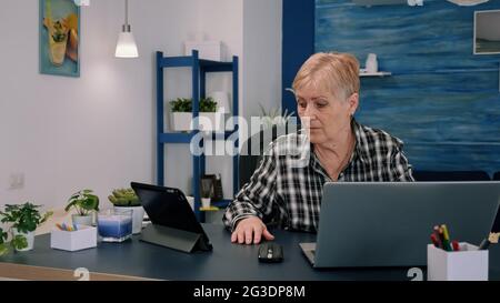 Mature woman using tablet and laptop in same time analysing financial graphs working from home sitting in workplace. Busy focused old employee writing, searching, analysing reports Stock Photo