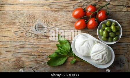 Two buffalo mozzarella, basil, cherry tomatoes and green olives on old wooden background, top view, space for text. Stock Photo