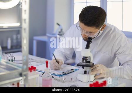 Scientist taking notes sitting at table with microscope in microbiology laboratory Stock Photo