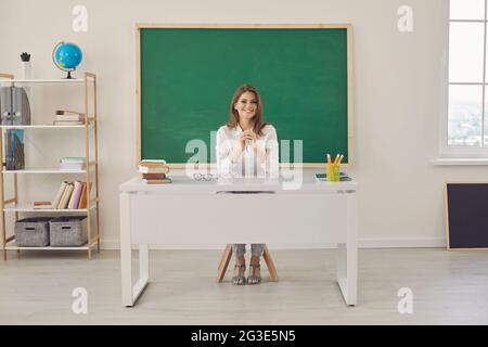 Young positive teacher sitting at the table teaches at a lesson in a school class. Stock Photo