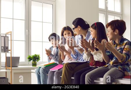 Female teacher together with group of happy children clapping hands while sitting in classroom. Stock Photo