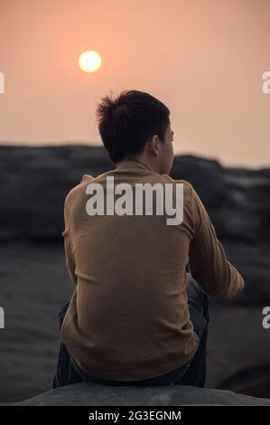 Loneliness young asian man sitting and looking at something on rock cliff at the sunset Stock Photo