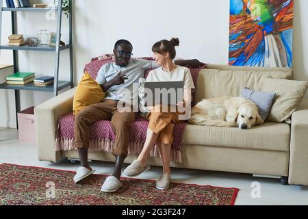 Young couple sitting on sofa with laptop and talking to each other in the room at home Stock Photo
