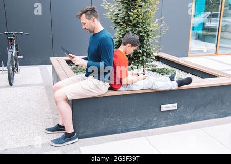 Father and son spend time in the city park, sitting back to back and using a smartphone or tablet.Modern technologies. Modern lifestyle Stock Photo