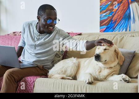 Young smiling man caring for his dog while sitting on sofa and working on laptop Stock Photo