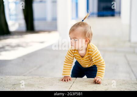 Little girl crawls on tiles in the courtyard of the house against the background of a lattice metal gate Stock Photo