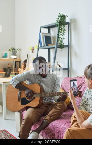 Young woman photographing young man on mobile phone while he playing guitar at home Stock Photo