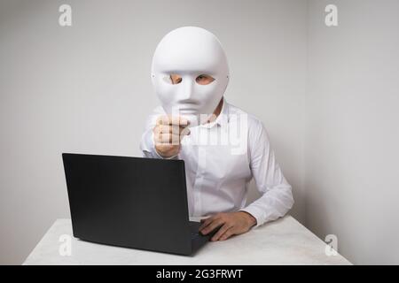 A man working at a laptop, covers his face with a mask, the concept of anonymity on the Internet Stock Photo