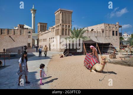 A mother and small daughter looking at a camel and camel owner in the old quarter of Dubai, the UAE., with house with a cooling wind tower just beyond Stock Photo