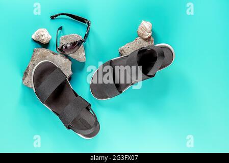 Summer holiday flat lay with women's accessories: black sandals, sunglasses, and seashells on blue background. Vacation and travel items. Stock Photo