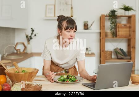 Young woman eating vegetable salad and using laptop in the kitchen Stock Photo