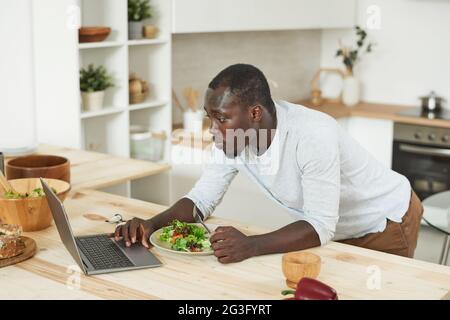 Young man eating vegetable salad and using laptop in the kitchen Stock Photo