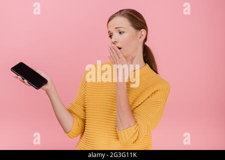 A surprised scared and amazed attractive woman in yellow sweater with broken phone screen in her hand set, looking to smartphone hand near face standi Stock Photo