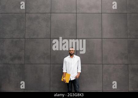 Focused serious young Afro American businessman, student or freelancer standing with yellow laptop outdoors, preparing for online meeting remotely Stock Photo
