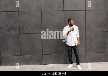 Full length portrait of pensive young Afro American millennial businessman or student, standing isolated outdoors in casual smart clothes, drinking Stock Photo