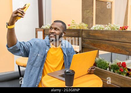 Handsome afro american man in trendy casual wear with yellow laptop in hand sitting in urban cafe outdoors, drinking take away coffee,hold remote talk Stock Photo