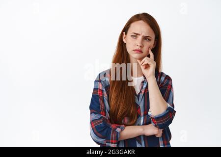Perplexed redhead girl student, look thoughtful or puzzled, frowning while thinking, making tough decision, standing against white background and Stock Photo