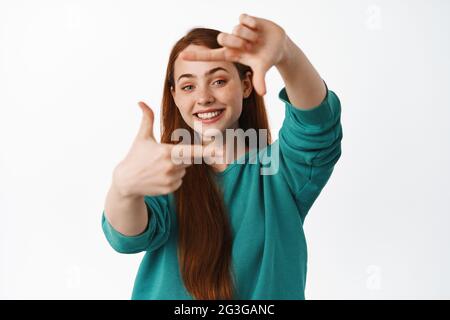 People and lifestyle. Happy redhead woman look through hand frames and smiling, imaging and getting creative, standing over white background Stock Photo