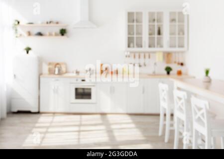 Blurred background of modern sunny kitchen. White kitchen furniture with utensils, plants in pots on shelves Stock Photo