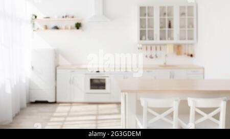 Sunny kitchen interior in Scandinavian style with white kitchen furniture and dining area, blurred background Stock Photo