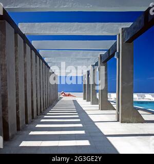 Historical 1981archive view of shadows & lead in lines from repetitive symmetrical columns and beams in1980s pergola beside new swimming pool woman sun bathing in part of a holiday refurbishment project of abandoned British Army officers homes close to the Maltese Pembroke Barracks an 80s archival image of a blue sky day on the main island of Malta in the Mediterranean Stock Photo