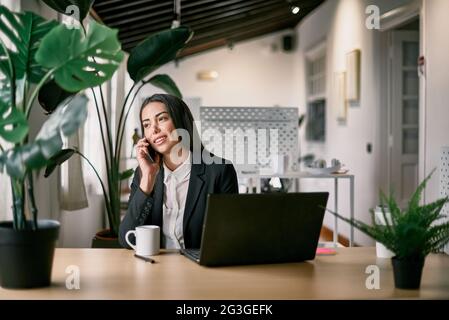 Smiling employee speaking on smartphone at desk with laptop Stock Photo