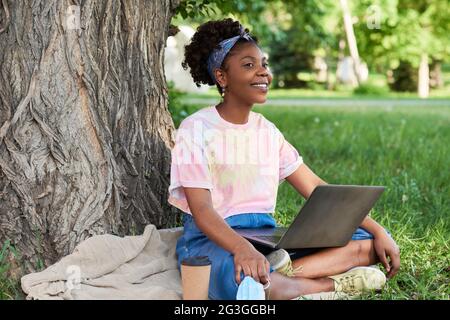 African businesswoman working on laptop while sitting on the grass in the park and drinking coffee Stock Photo