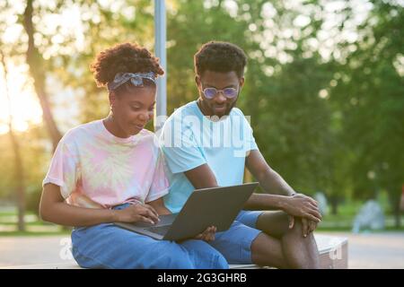 Two friends sitting on the bench nd using laptop together in the park Stock Photo