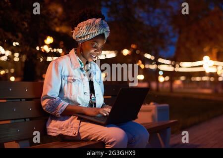African young woman sitting on bench and using laptop in the park in the evening Stock Photo