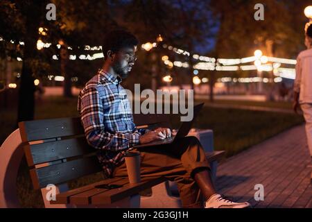 African young man sitting on bench and using laptop in the park in the evening Stock Photo