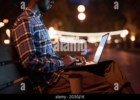 Young man sitting on the bench and working online on laptop in the park in the evening Stock Photo