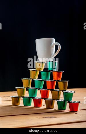 Cup of coffee on top of a tower made with colored coffee pods and used on wooden table on black background. Stock Photo