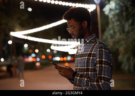 African young man reading a message online on mobile phone while standing in the city in the evening Stock Photo