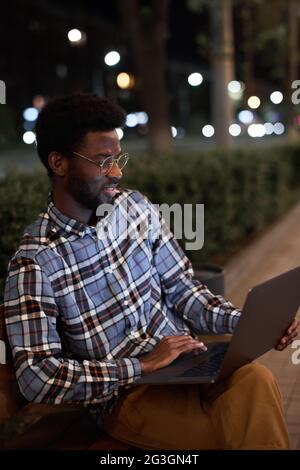 African young man sitting on the bench and working online using his laptop while sitting in the evening in the park Stock Photo