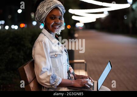 African young woman smiling at camera while sitting on the bench and working online on laptop outdoors Stock Photo