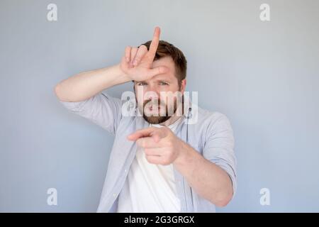 Annoyed Caucasian man in casual clothes insults someone with a loser gesture, fingers on forehead and viciously points forefinger. Stock Photo