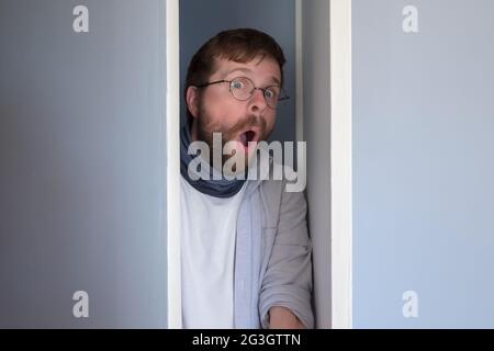 Bearded man in casual clothes is hiding in a closet, he peeps through the open door and looks in surprise with an open mouth and big eyes.  Stock Photo