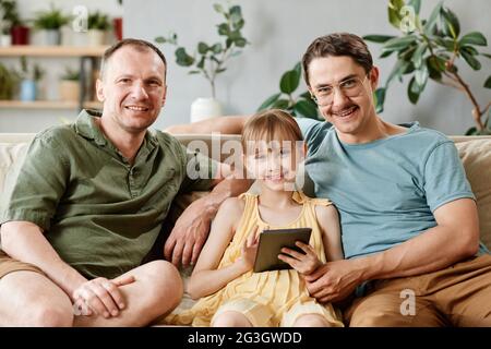 Portrait of gay family sitting on sofa with adopted daughter using digital tablet at home Stock Photo