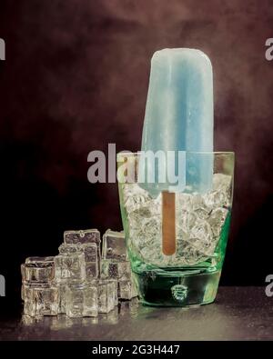 A blue popsicle is standing inside a glass with ice and smoke on the dark background Stock Photo