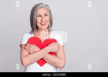 Photo of dreamy sweet age woman dressed white outfit embracing large red heart looking empty space isolated grey color background Stock Photo
