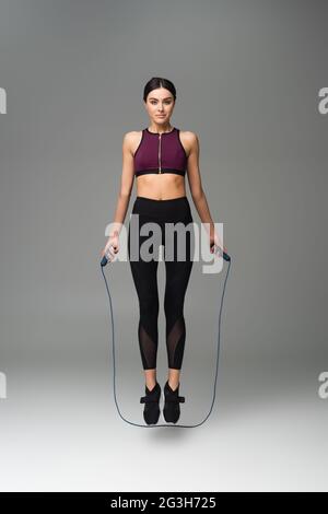 sportswoman looking at camera while jumping with skipping rope on grey background Stock Photo