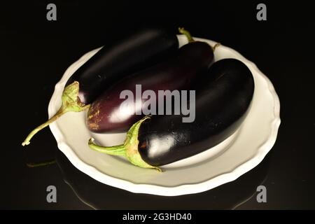 Three organic dark eggplants on a white dish, close-up, on a black background. Stock Photo