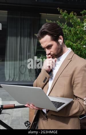 pensive businessman in earphones holding laptop near building outside Stock Photo