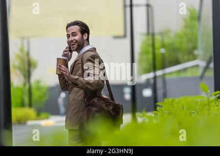 cheerful businessman in formal wear talking on smartphone and holding paper cup outside Stock Photo