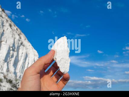 Man hand is holding piece of chalk.. Chalk mountains against the blue sky in the day. Natural Chalk mining. Stock Photo