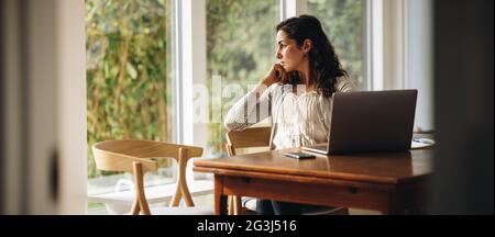 Woman sitting at table with laptop looking away. Woman taking a break from work at home. Stock Photo
