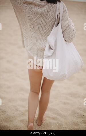 Beautiful stylish woman with tote bag walking on sandy beach to sea, carefree moment. Stylish young female in knitted sweater enjoying vacation and re Stock Photo