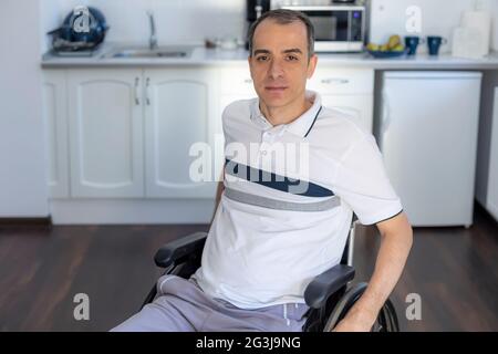 Smiling Young Handicapped Man Sitting On Wheelchair In Kitchen.  Young man infront of kitchen. Focus on his face. Stock Photo