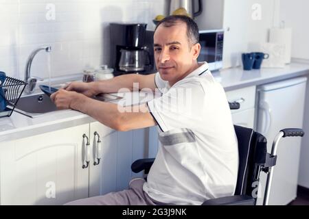 Young man Disabled Man On Wheelchair Washing Dishes. Smiling Young Handicapped Man Sitting On Wheelchair In Kitchen. Focus on his face. Stock Photo